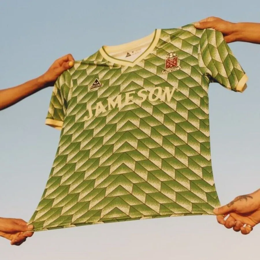 Hands holding up a green and white soccer jersey featuring a repeating 3D geometric chevron pattern, a V-neck collar