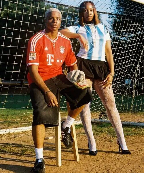 A young man sitting on a wooden stool and a woman standing next to him in front of a soccer goal net. They are wearing a red sports jersey and a light blue striped jersey respectively.