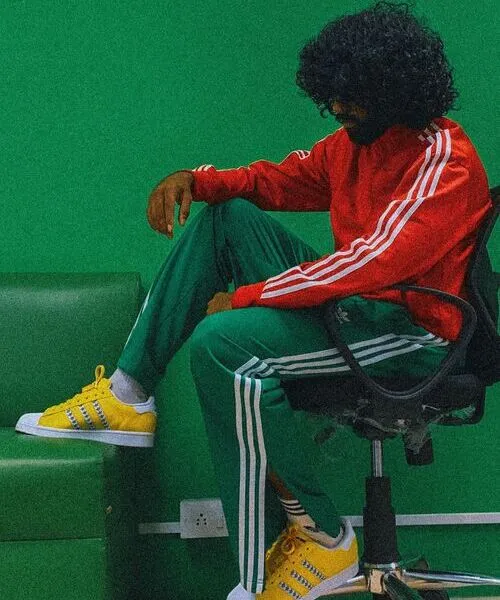 A person with curly hair sitting in an office chair against a green wall, wearing a bright red zip-up sports jacket and green track pants with white stripes.