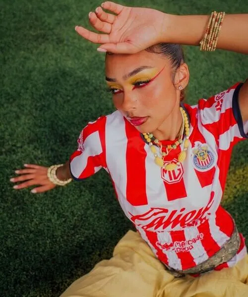 Close-up of a young woman posing on a green turf field, wearing a red and white striped football jersey with vibrant yellow and red eye makeup. She is styled with beaded jewelry, including necklaces and bracelets, blending sports apparel with high-fashion aesthetics.