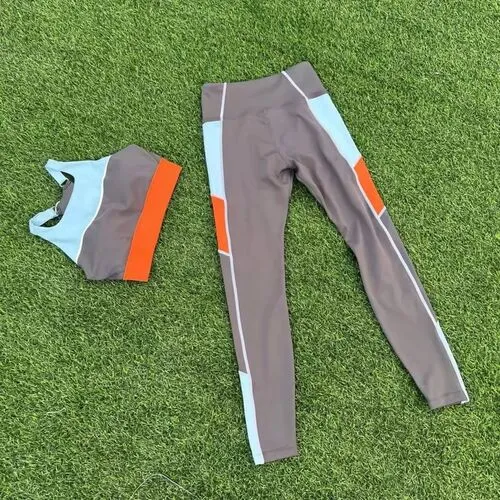 A two-piece women's athletic set laid out on green grass, including a racerback sports bra and matching high-waisted leggings in a color-block design of charcoal grey, light blue, and vibrant orange with white piping.