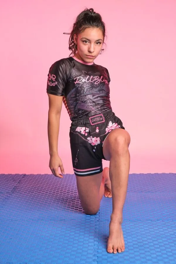 A young woman kneeling on a blue mat against a pink background, wearing a black short-sleeved compression shirt and matching shorts featuring pink floral patterns and stylized lettering.