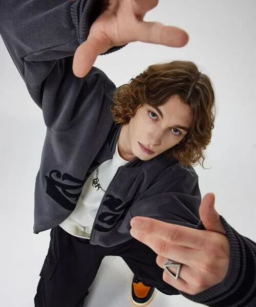 A young man with wavy hair posing in a dynamic top-down view, wearing a charcoal grey fleece zip-up jacket over a white t-shirt and black cargo pants.