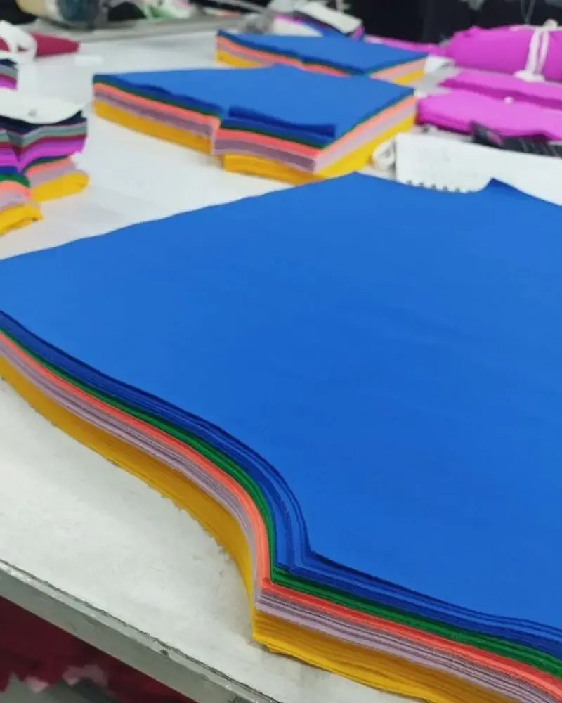 A close-up view of neatly stacked layers of multi-colored fabric, including bright blue, yellow, and pink, precisely cut into garment patterns on a habit fit sports factory production table.