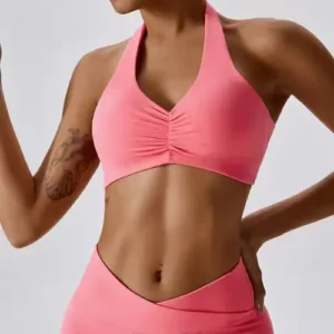 A woman wearing a coral pink halter-style sports bra with a gathered cinched front detail and a V-shaped waistband.