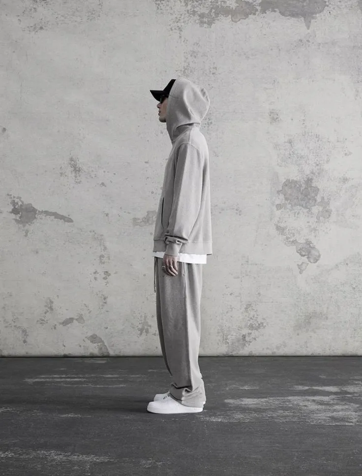 A side profile shot of a person standing against a weathered concrete wall, wearing a light grey oversized hoodie with the hood up, matching wide-leg trackpants, a black cap.