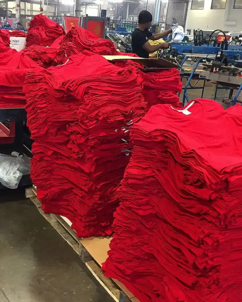 Large stacks of folded red t-shirts piled high on wooden pallets inside a clothing factory, with a worker operating machinery in the background.