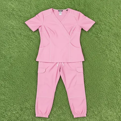 A complete set of light pink medical scrubs laid out on green grass, including a short-sleeved V-neck wrap-style top and matching jogger-style pants with side cargo pockets.