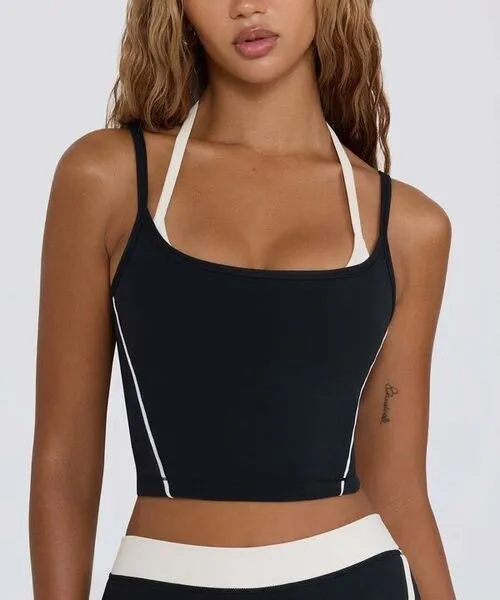 A woman wearing a dual-layered black athletic crop top featuring a white halter-neck inner layer and a black square-neck outer layer with thin white vertical piping; paired with matching black and white high-waisted leggings.