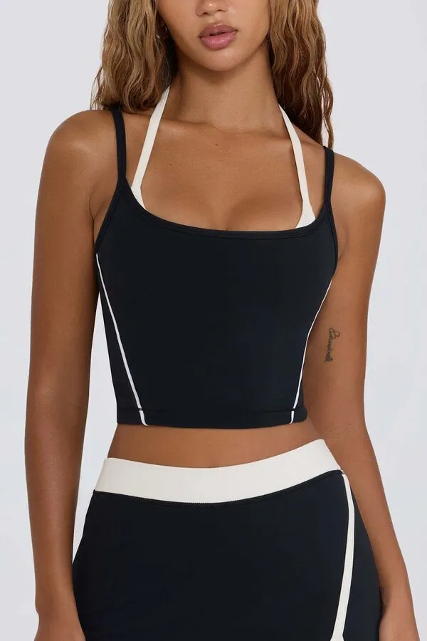 A woman wearing a dual-layered black athletic crop top featuring a white halter-neck inner layer and a black square-neck outer layer with thin white vertical piping; paired with matching black and white high-waisted leggings.