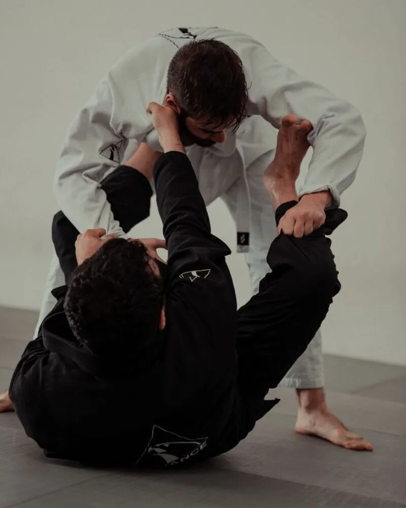 Two martial artists engaged in a Brazilian Jiu-Jitsu (BJJ) sparring session on a mat; one athlete is wearing a traditional white pearl weave Gi while the other is in a black reinforced Gi, demonstrating high-level grappling techniques and the durability of professional-grade training uniforms.
