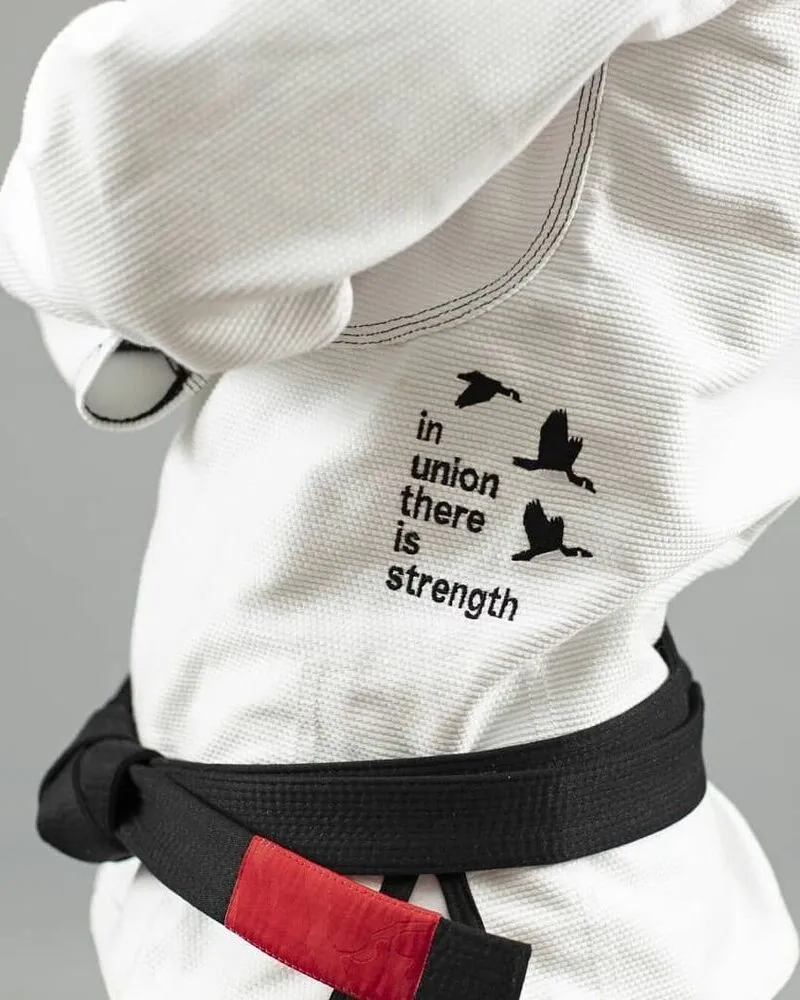 A close-up of a high-quality white Brazilian Jiu-Jitsu (BJJ) Gi featuring a heavy pearl weave fabric texture; it includes a black custom embroidery of flying birds with the quote "in union there is strength" on the side, paired with a professional black belt with a red rank bar.