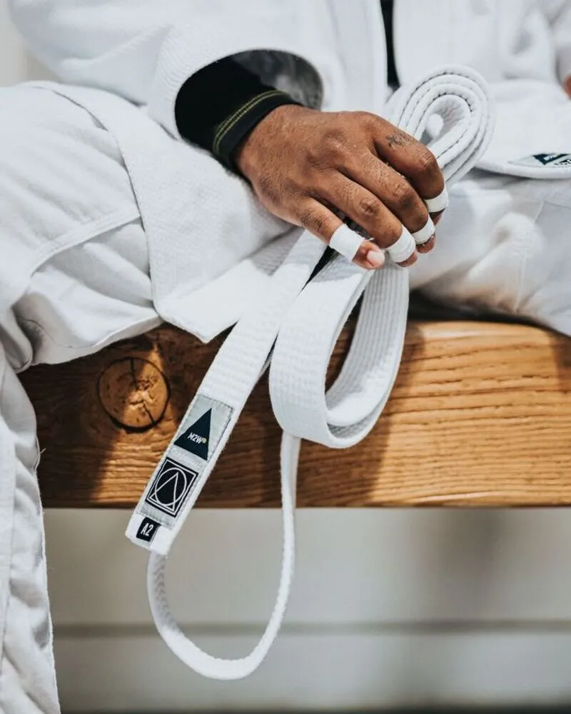 A close-up shot of a martial artist sitting on a wooden bench, holding a neatly folded white BJJ belt with a black rank sleeve; the image highlights the reinforced stitching of the white Gi sleeves, taped fingers for grip protection, and a custom "A2" size and brand woven label on the belt.