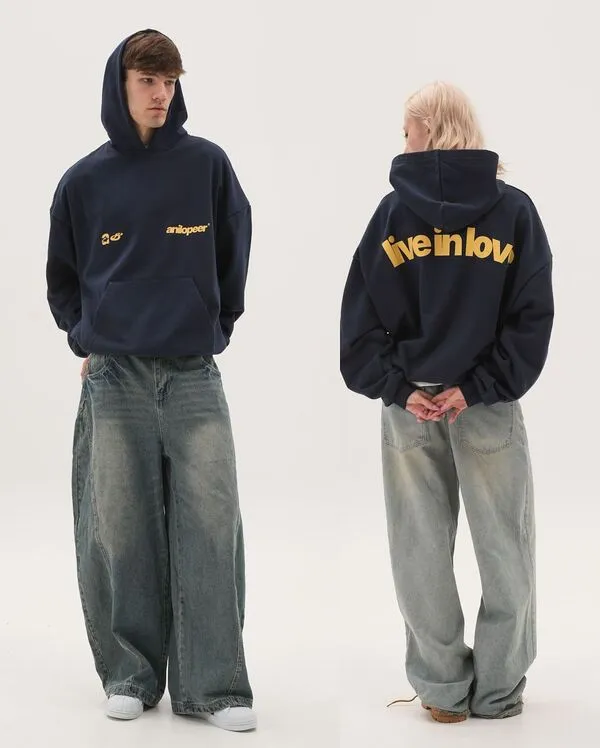 A front and back view of a navy blue oversized streetwear hoodie featuring yellow "anilopeer" branding on the chest and a bold "live in love" slogan on the back; paired with extra-wide baggy acid-wash denim jeans and white sneakers for a modern urban aesthetic.